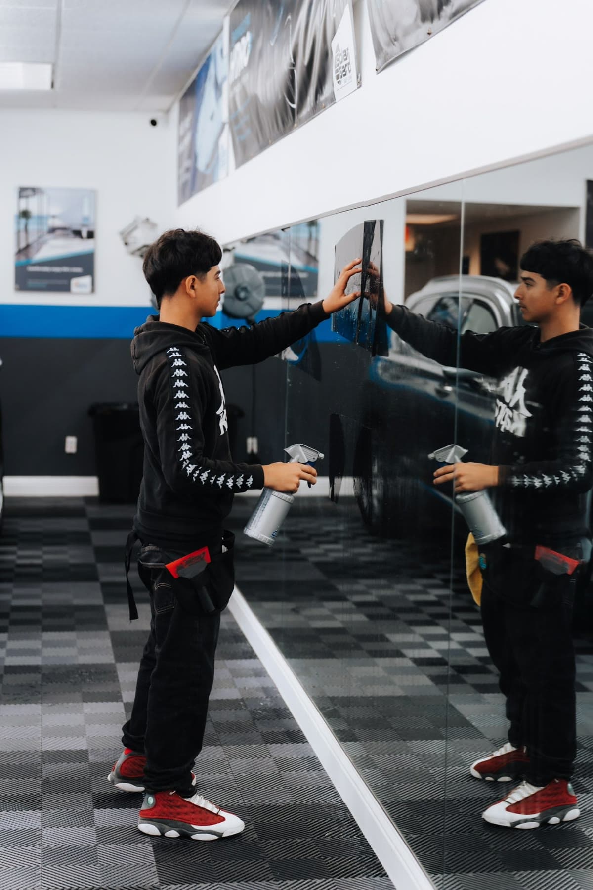 A student in a black hoodie holding a sheet of dark window film against a tall mirrored glass wall with one hand and a spray bottle in the other, his reflection beside him in the training shop