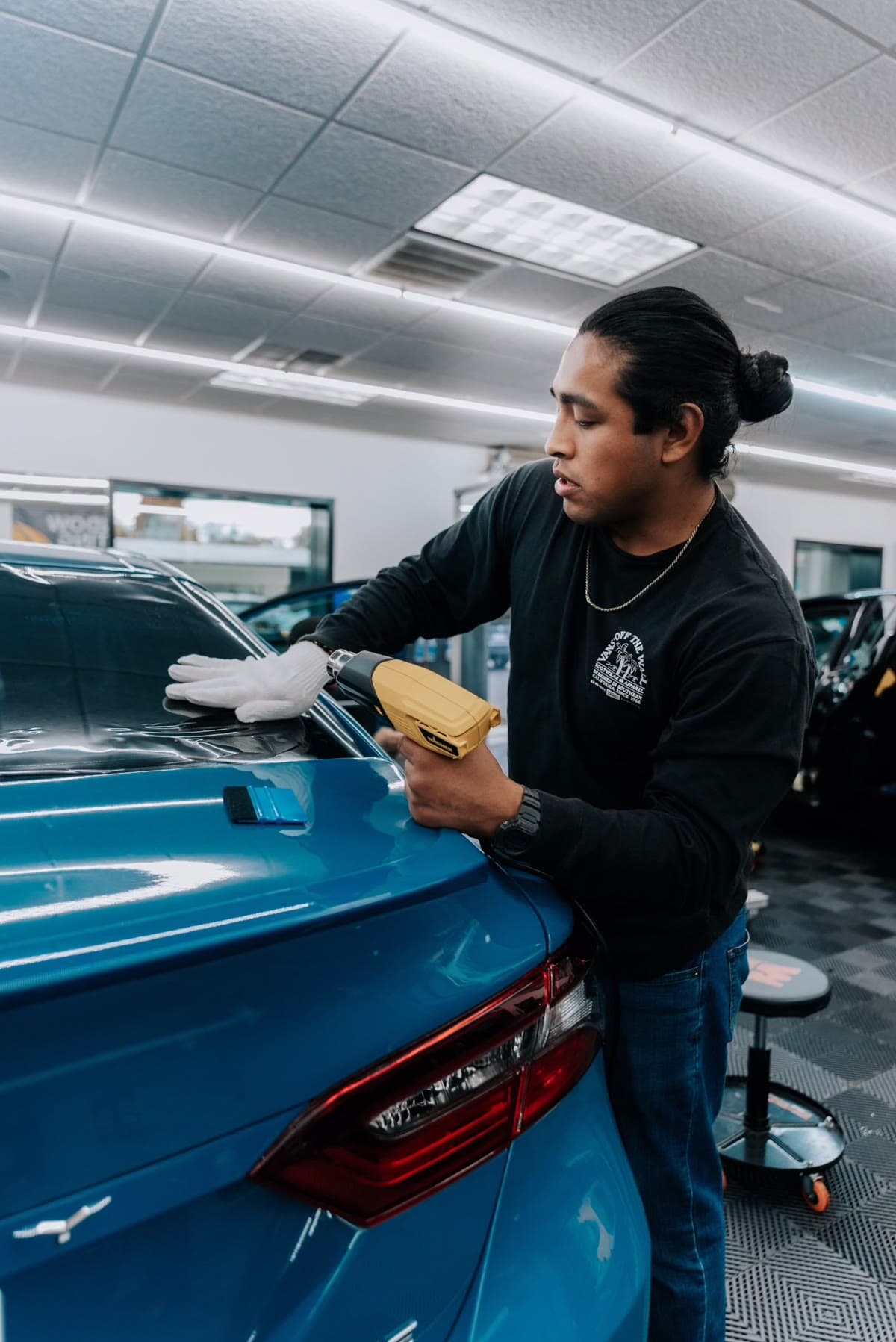 A student in a black long-sleeve shirt shrinking dark window film onto the rear windshield of a bright blue sedan with a heat gun, one gloved hand pressing the film flat, in the Modesto training shop