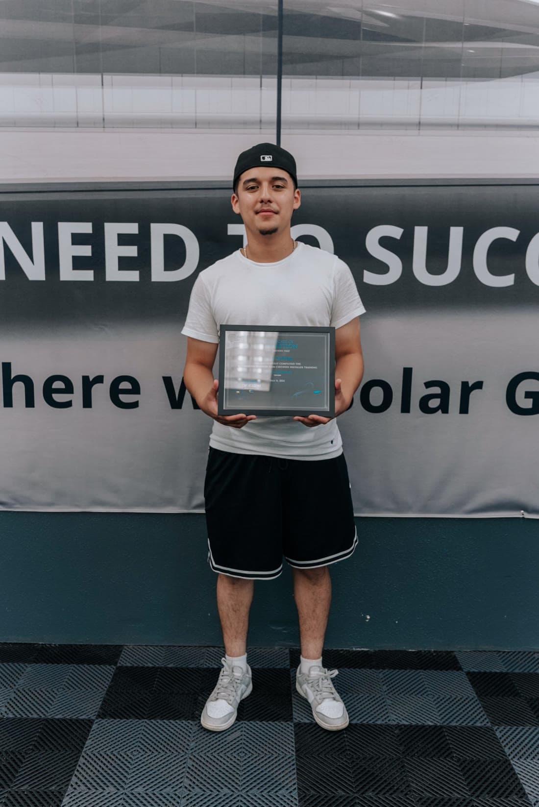 Un graduado con playera blanca y gorra sosteniendo su certificado de finalización enmarcado de California Tint Academy frente a la pared de Solar Gard