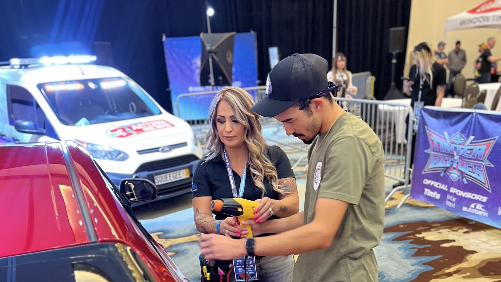 Roberta Rodríguez sosteniendo una pistola de calor junto a un competidor, al lado de un carro rojo en el piso de Tinter Battles, con el letrero de patrocinadores del evento detrás.